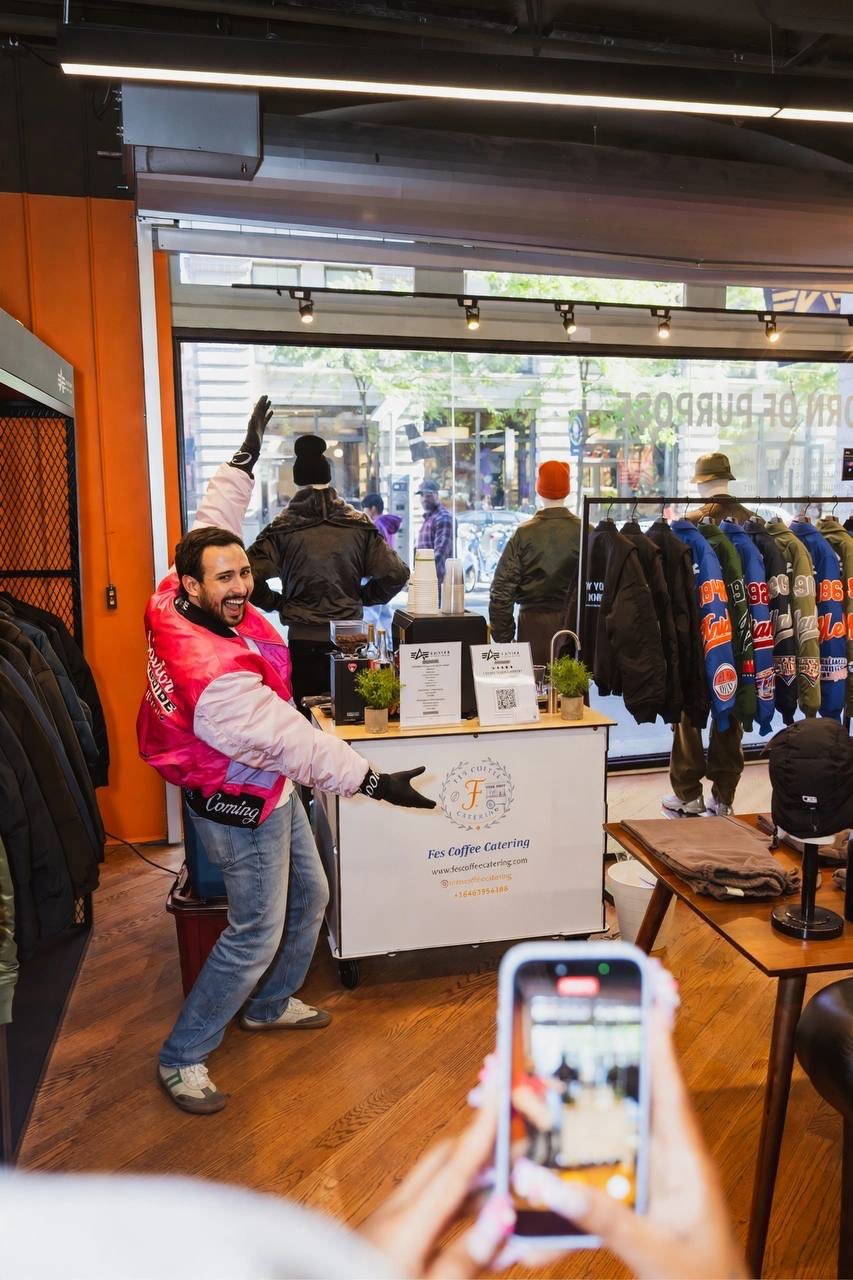 A barista at the Fes Coffee cart in a clothing store, smiling as a guest films him with a phone