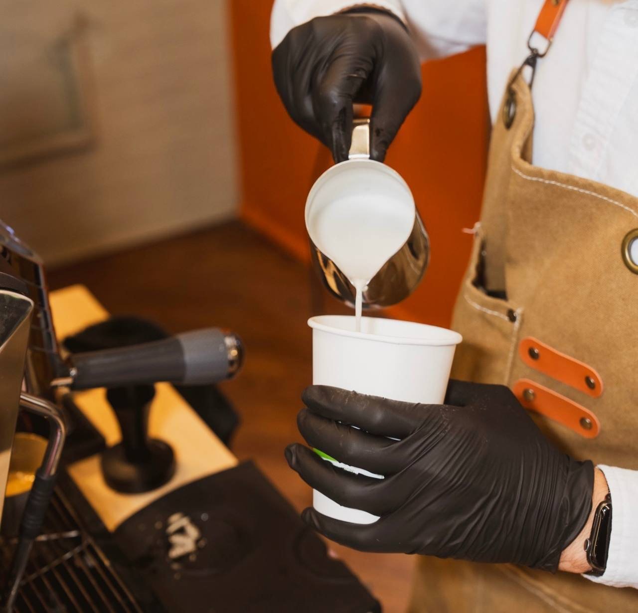 Barista in black gloves pouring steamed milk into a paper cup at the espresso machine