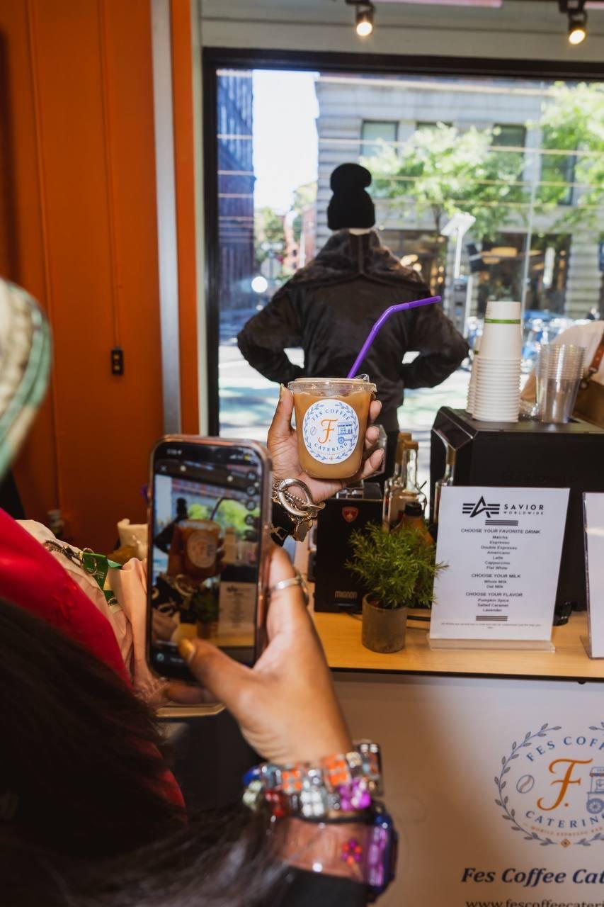 Guest holding up an iced latte in a Fes Coffee cup in front of the cart at a store event