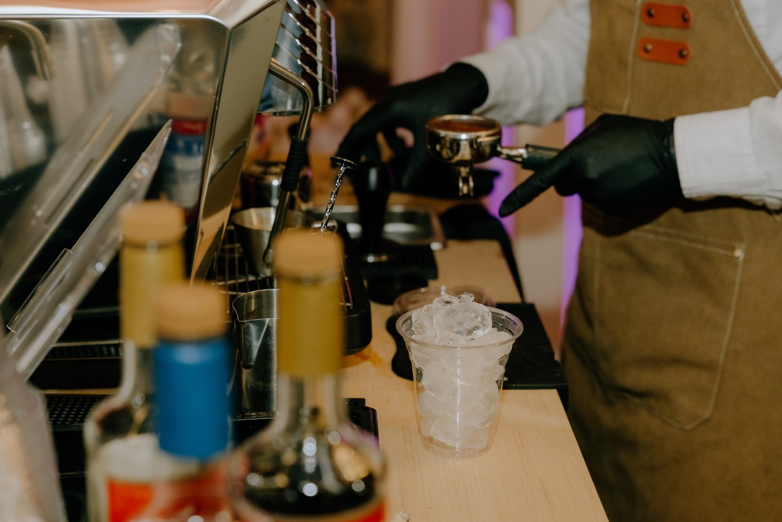 Barista in black gloves at the espresso machine with syrup bottles and an iced cup on the counter